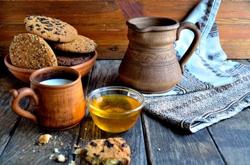 Oatmeal cookies in a ceramic rustic plate, milk in a ceramic mug, napkin, honey on a rustic wooden table.
