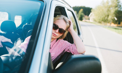 Young caucasian blond woman traveling by car and having fun on her summer holiday journey. Happy girl driving car on countryside road.