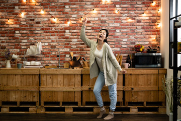 Happy woman with eyes closed carefree raise hand dancing singing in kitchen while cooking. beautiful relaxed lady in warm house indoor winter with red brick wall and light decorated in background