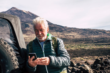 Caucasian old man use modern technology cellular phone at the mountain during trip vacation with off road car at the mountain - planning maps and roads enjoying the nature outdoor for vacation