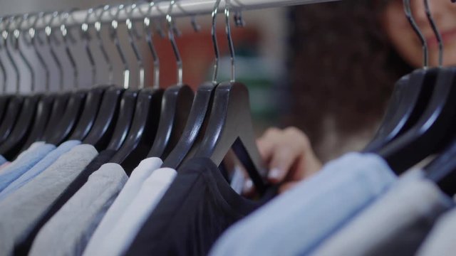 Shopper is moving hangers with tee shirts in sportive clothing store, close-up