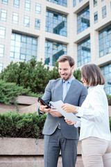 Handsome young male and female business people talking in front of an office building, having a meeting and discussing, comparing data on tablet and paper reports. Work anywhere concept.