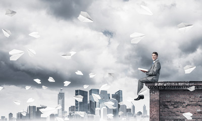 Man on roof edge reading book and cityscape at background