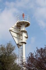 Lighthouse beacon warning light on top of strong metal structure surrounded with safety fence and tall trees on cloudy blue sky background