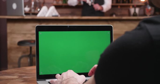 Close up shot Businessman typing fast on a computer with green screen in a vintage pub or restaurant. A bartender sits at the bar counter in the background