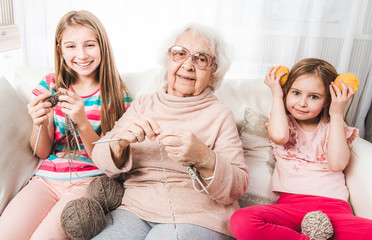 Smiling granddaughters with grandmother knitting together - obrazy, fototapety, plakaty