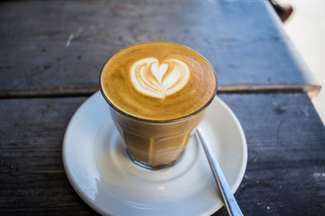 Thick glass tumbler with cappuccino and latte-art in the form heart. Ffresh coffee with milk. On a wooden table with saucer, spoon
