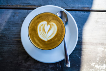 Thick glass tumbler with cappuccino and latte-art in the form heart. Ffresh coffee with milk. On a wooden table with saucer, spoon