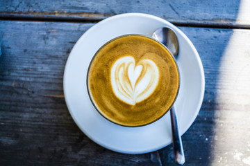 Thick glass tumbler with cappuccino and latte-art in the form heart. Ffresh coffee with milk. On a wooden table with saucer, spoon