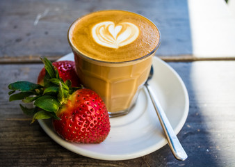 Thick glass tumbler with cappuccino and latte-art in the form heart. Fresh strawberry and coffee. On wood with saucer, spoon.