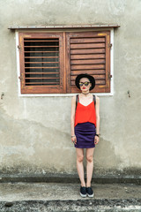 A girl in a black hat stands near the window with wooden shutters on the background of a concrete wall