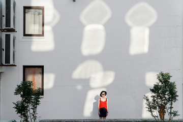 Summer girl in a black hat and sunglasses stands near a white wall with reflections from the sun reflected from windows