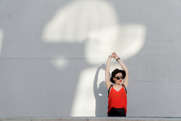 Summer girl in a black hat and sunglasses stands near a white wall with reflections from the sun reflected from windows