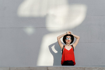 Summer girl in a black hat and sunglasses stands near a white wall with reflections from the sun reflected from windows
