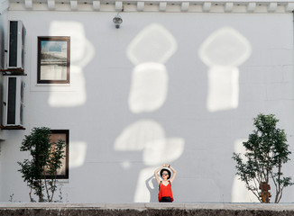 Summer girl in a black hat and sunglasses stands near a white wall with reflections from the sun reflected from windows