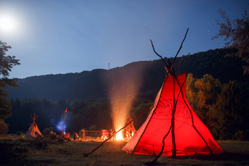 Image of camping with red tipi and people near bonfire on dark forest background. beautiful lanscape background. © Dragosh