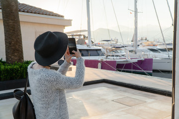 A girl in sunglasses and a black hat photographs on a smartphone a yacht in the harbor in a European resort town looking into the distance. Shot from the back