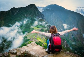 View of girl sitting on the edge of the rock with raised hands and taking pleasure of breathtaking forested mountains landscape of Machupicchu