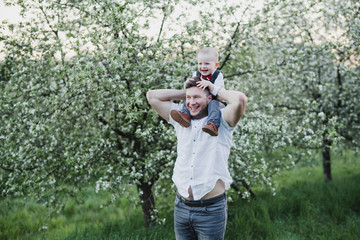 Happy stylish caucasian father and son walking and spending time together in blooming spring garden. Dad playing with child outdoors. Love, parenthood, childhood, happiness concept.