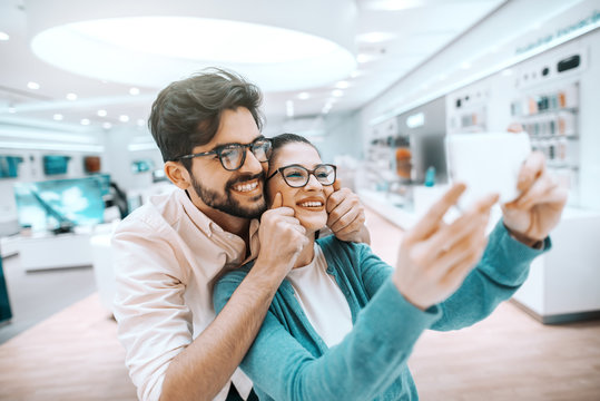 Cheerful multicultural couple with eyeglasses dressed casual having fun at tech store and taking selfie.