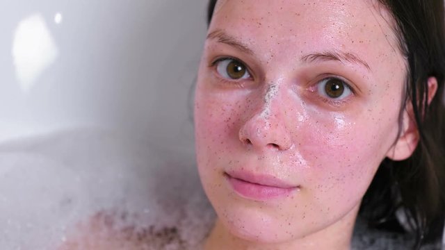 Beautiful brunette woman with scrub on the face lying in the bathroom at home and smiling. Face close-up.