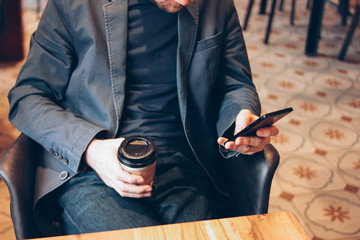 Adult man drinking coffee from paper cup and using mobile phone at cafe