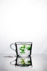 Green color dissolves in a cup of water on a white background.The mug is reflected on the wet surface. At the top there is space for text.