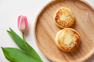Closeup fresh flower pink tulip on wooden round plate, two golden fried cheesecakes in the shape of a figure 8, dessert made from cottage cheese. Concept tasty and healthy breakfast March 8