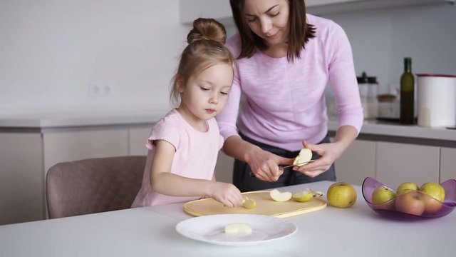 Mom and daughters cooking healthy food at home and having fun. Young woman cutting yellow apple on the desk in front her daughter. Household, teamwork helping, maternity concept. Slow motion