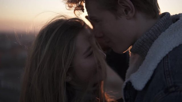 Close up footage of romantic young couple standing face to face backlit by the sun with flare effect while standing on the roof in wind with an urban background