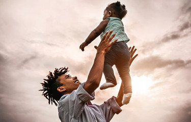 Happy father playing with his baby daughter during sunset time - Afro family having fun outdoor -...