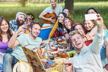 Group of diverse friends taking photo selfie while doing picnic in park - Happy young people having with new mobile phone technology and eating outdoor - Millennial, friendship, food and drink concept