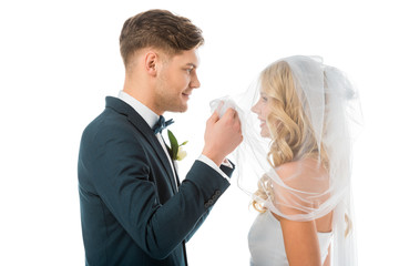 happy groom looking at smiling brides face while lifting bridal veil isolated on white