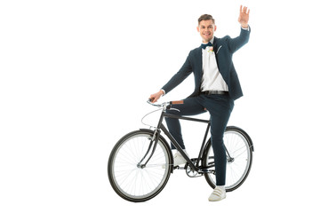 Fototapeta na wymiar cheerful groom in elegant suit and sneakers sitting on bike and waving hand isolated on white