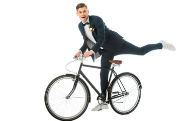 cheerful groom in elegant suit making stunts on bike isolated on white