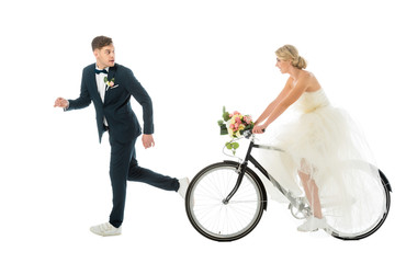 handsome groom in suit and sneakers running from bride on bicycle isolated on white