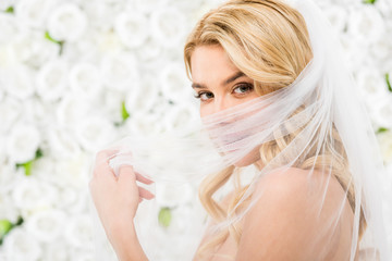 beautiful young bride hiding face behind bridal veil while looking at camera on white floral background