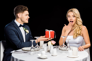 smiling young groom presenting red gift box to surprised bride isolated on black