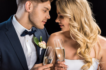 happy couple sitting face to face and clinking glasses of champagne isolated on black