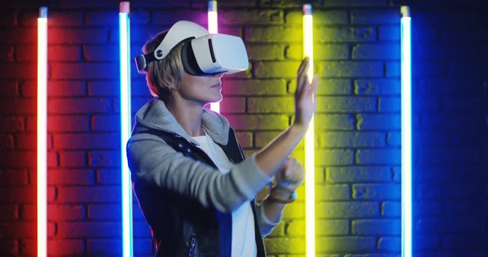 Young Caucasian woman with short fair hair in VR goggles playing a virtual game on the neon colorful lamps background.