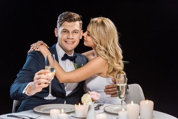happy young bride embracing handsome groom with glass of champagne isolated on black
