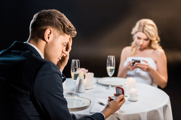 selective focus of sad young man holding gift box with wedding ring while girl using smartphone on black background