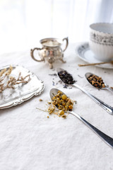 Different types of tea on vintage metal spoons on white fabric background, teacup, dried flower, silver plate, cookies. Vintage food and drink setting styling.  Organic healthy well-being lifestyle.