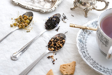 Different types of tea on vintage metal spoons on white fabric background, teacup, dried flower, silver plate, cookies. Vintage food and drink setting styling.  Organic healthy well-being lifestyle.