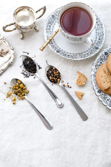 Obraz na płótnie Canvas Different types of tea on vintage metal spoons on white fabric background, teacup, dried flower, silver plate, cookies. Vintage food and drink setting styling. Organic healthy well-being lifestyle.