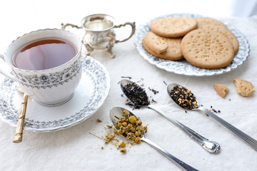 Different types of tea on vintage metal spoons on white fabric background, teacup, dried flower, silver plate, cookies. Vintage food and drink setting styling.  Organic healthy well-being lifestyle.