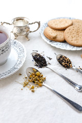 Different types of tea on vintage metal spoons on white fabric background, teacup, dried flower, silver plate, cookies. Vintage food and drink setting styling.  Organic healthy well-being lifestyle.