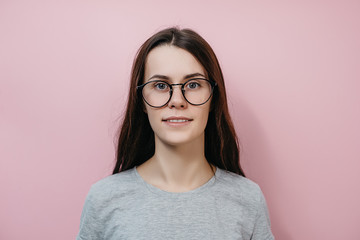 Portrait of charming young woman in transparent glasses smiling . Caucasian brunette female in gray T-shirt isolated on pink wall. People and emotion concept