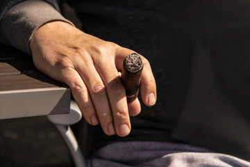 Close-up of man's hand resting on side of table beside his chair holding a lit cigar