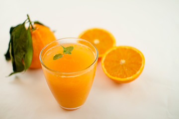 An inviting glass full of orange juice with a fresh mint leaf that floats. In the background an orange divided in two and a whole one with leaves on a white background